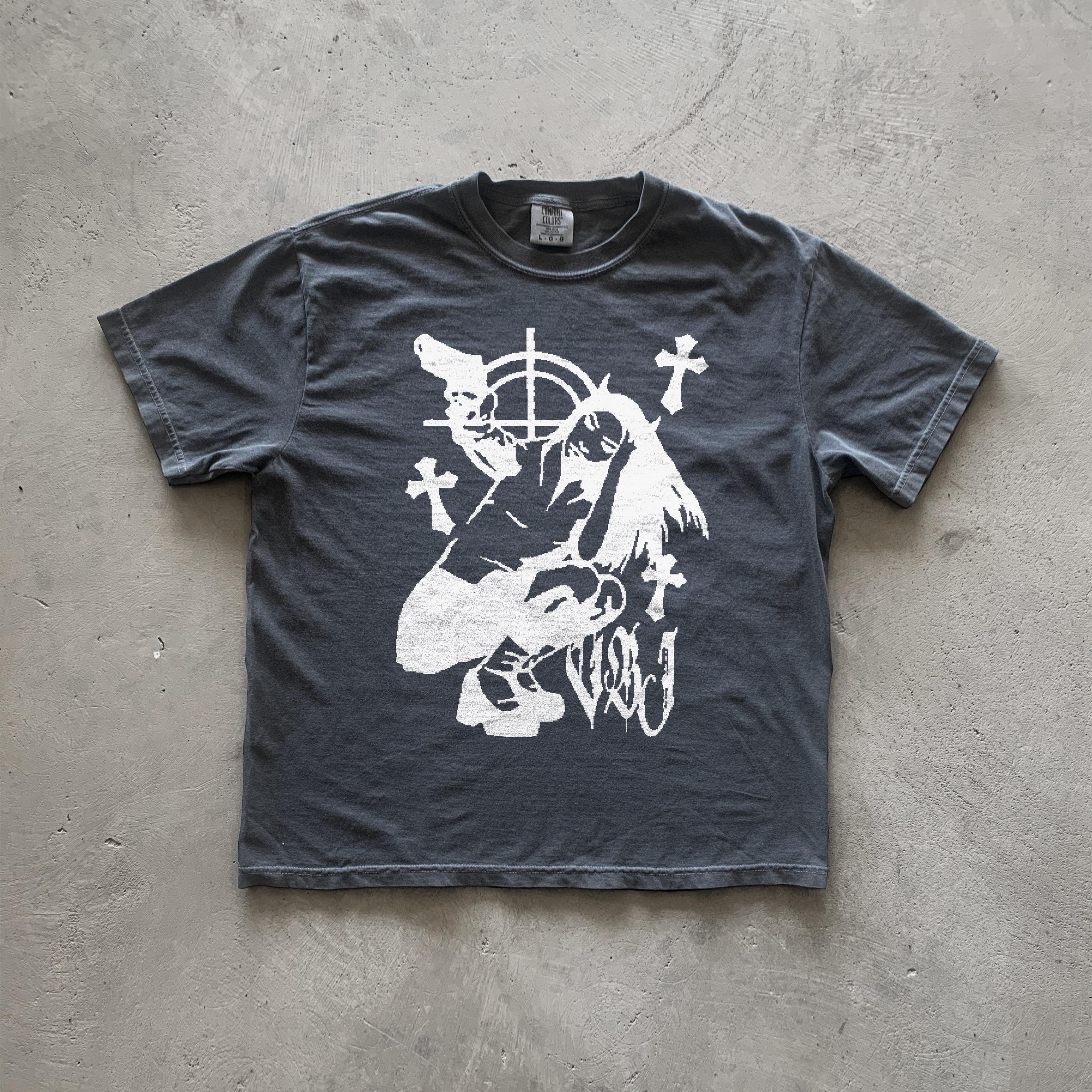 May include: Dark gray t-shirt featuring a graphic design of a stylized female figure holding guns, surrounded by crosses and a target symbol. The style is reminiscent of vintage punk or grunge aesthetics. The graphic is printed in white. The shirt appears to be made of a soft, comfortable fabric.