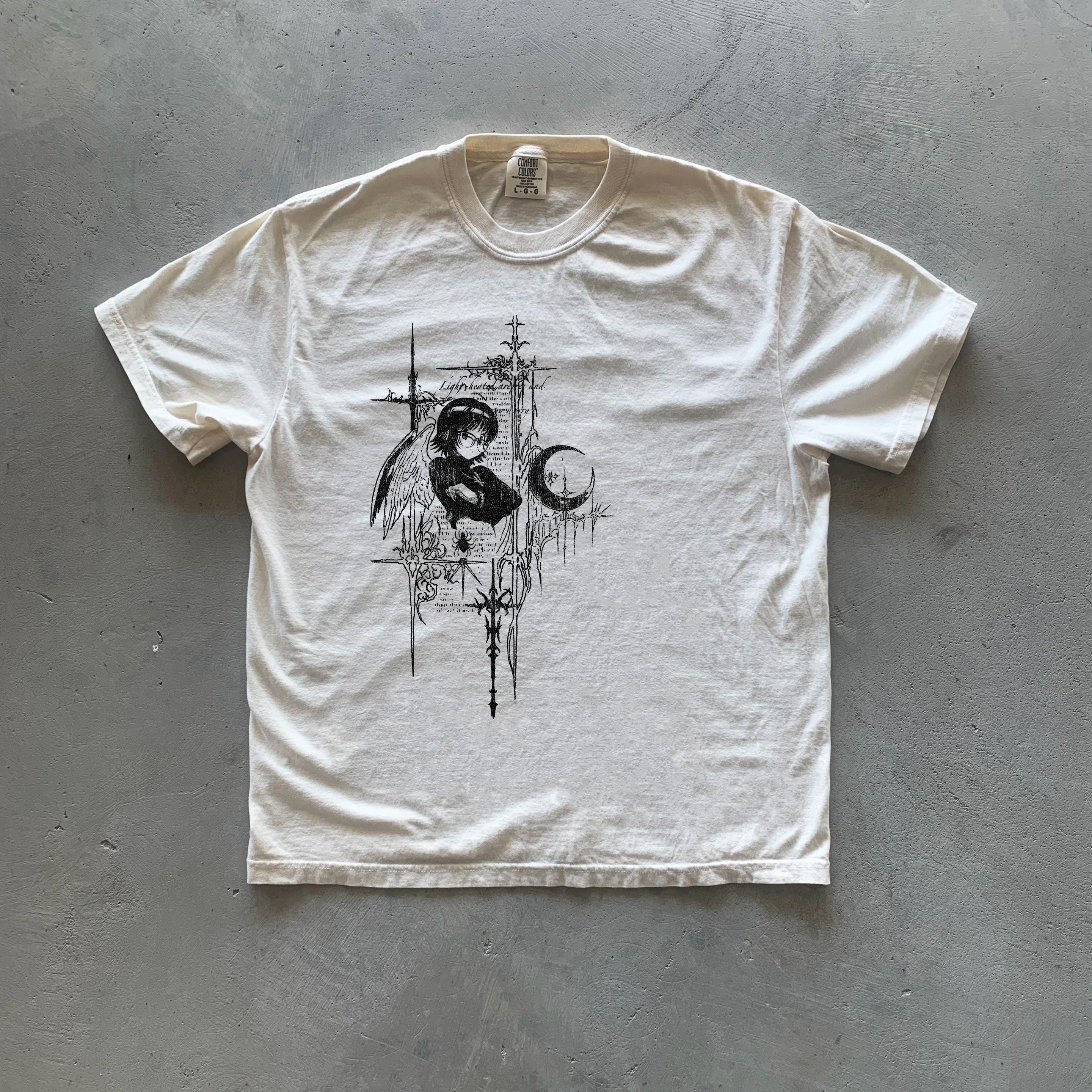 May include: A white t-shirt with a black and white graphic print of an angel with wings, a crescent moon, and a cross. The graphic is surrounded by a black and white design of stylized flowers and leaves.