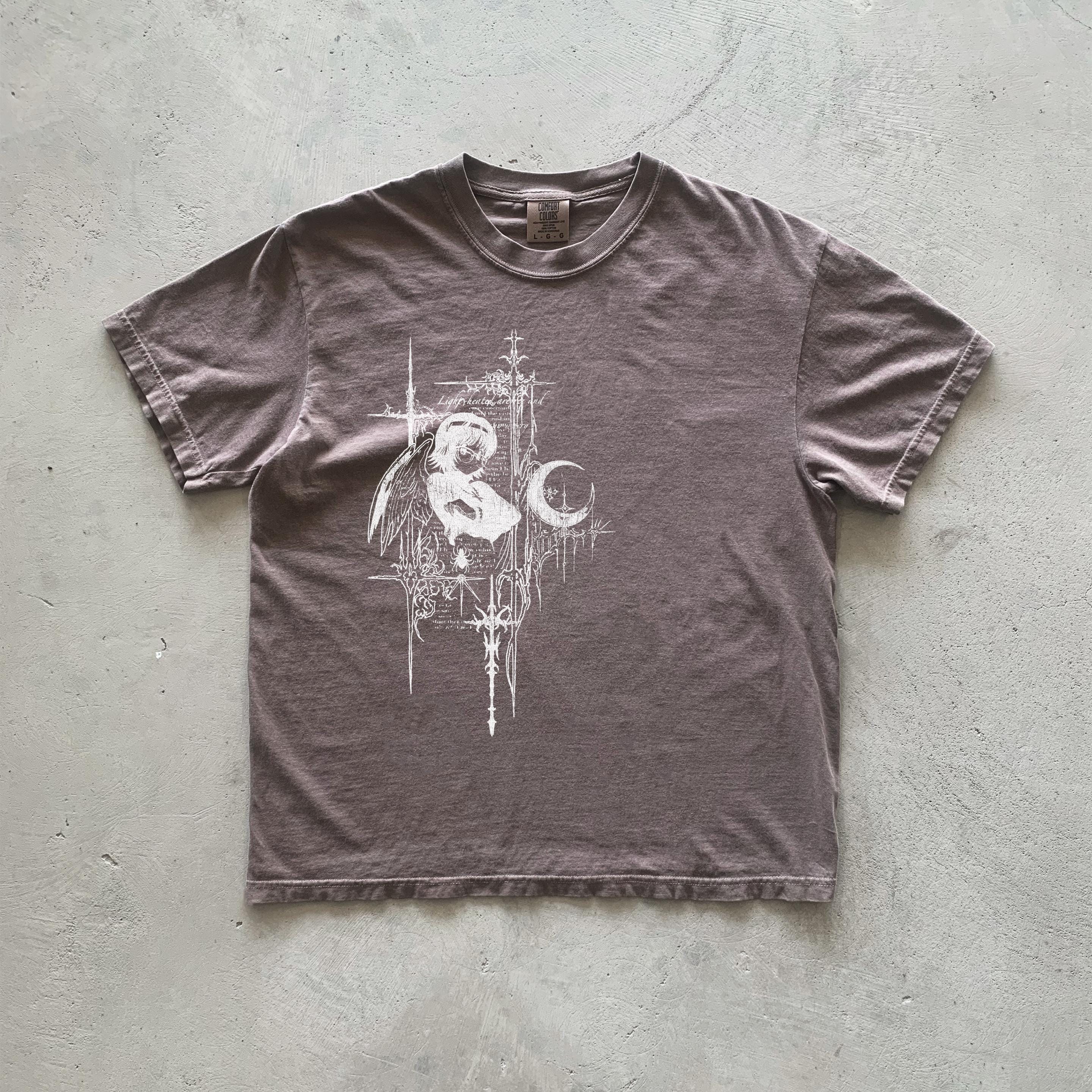 May include: A brown t-shirt with a white graphic print of an angel holding a crescent moon. The graphic is surrounded by a decorative border of lines and crosses.