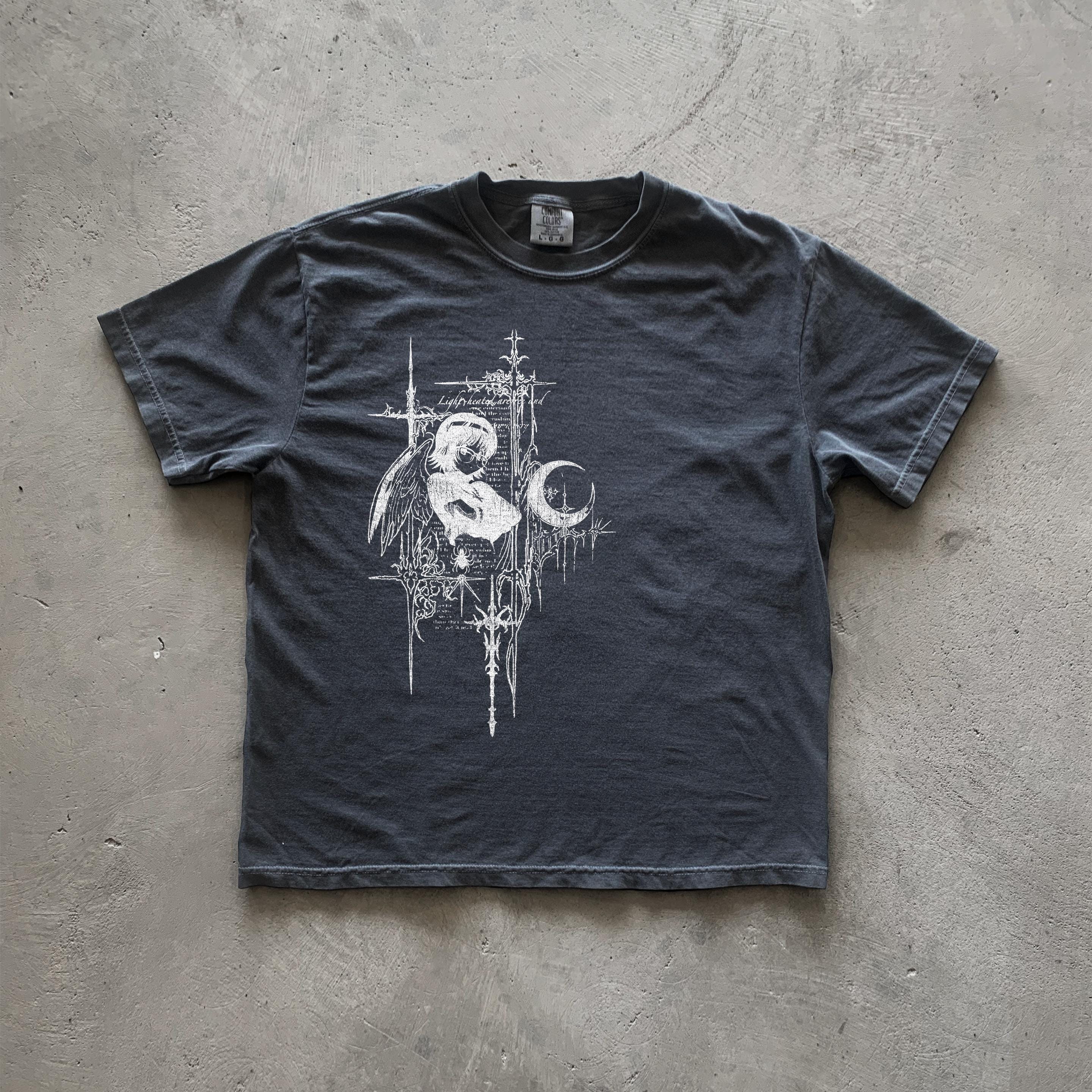 May include: A dark gray t-shirt with a white graphic print of an angel with wings, a crescent moon, and a cross. The graphic is surrounded by a decorative border of lines and crosses. The text 'Lightbringer' is included in the graphic.