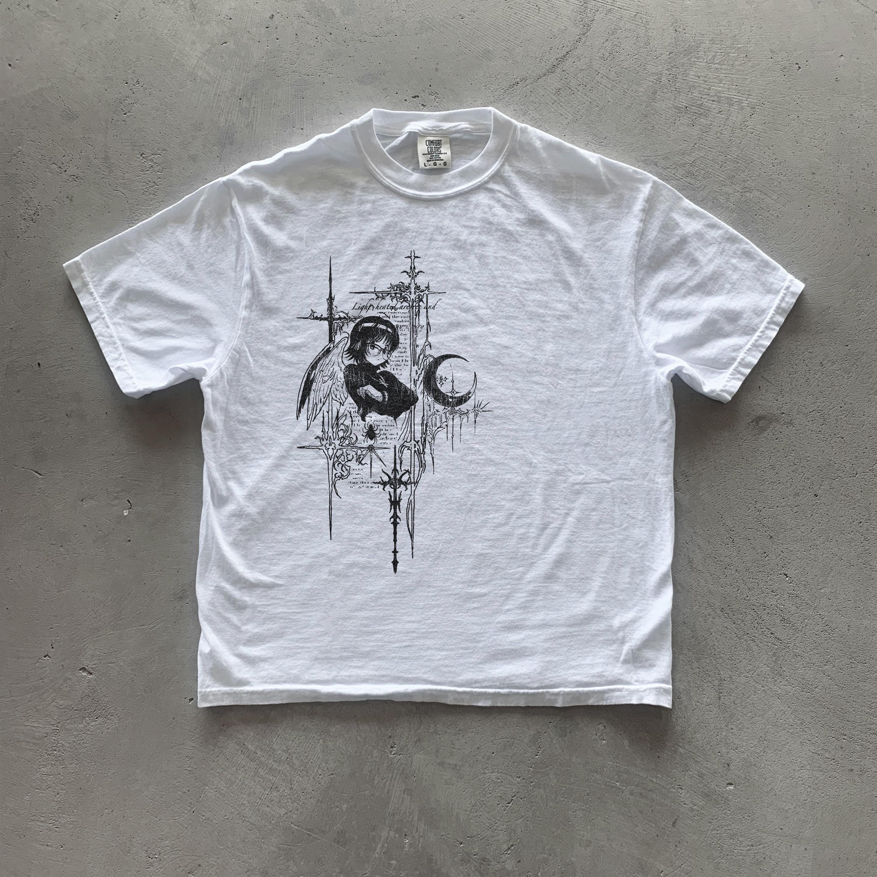 May include: White t-shirt with a black and white graphic print of a person with wings and a crescent moon. The graphic is surrounded by a design of crosses and other symbols. The text 'Light Within' is visible on the graphic.